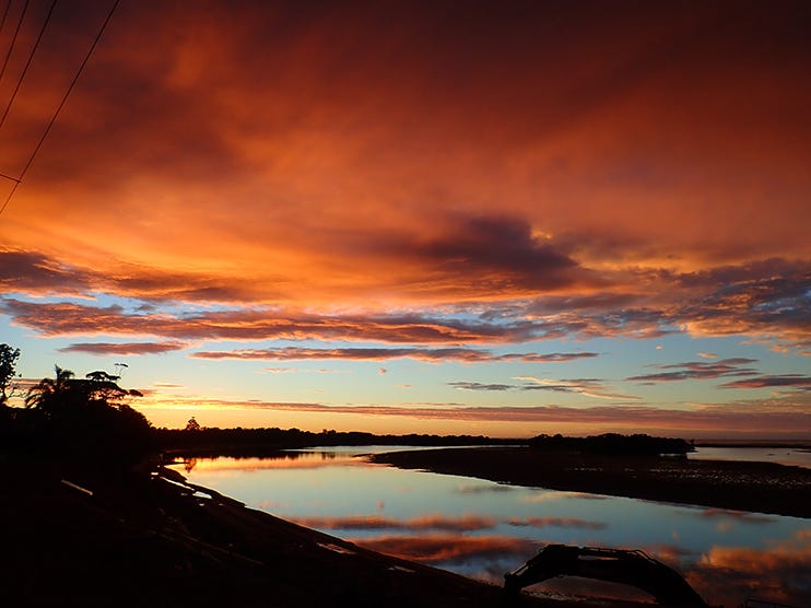 It’s a bright sunrise on a calm morning. A river with a glassy surface curves through the land that is quite dark. The horizon is lightly lit in a soft gold. There is a stretch of light blue sky, above which are scattered clouds and then a big bank of light clouds that are red-orange in color. Some of these clouds are reflected in the river.