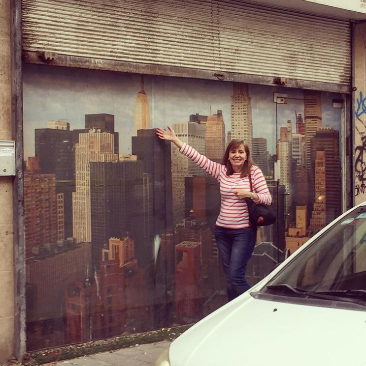 A woman in a striped shirt smiles at the camera and enthusiastically gestures to a wall mural of New York City’s skyline.