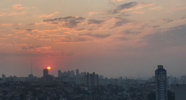 Imagem de um por-do-sol, sol alaranjado baixo no horizonte, com nuvens estreitas e esparças em cores de rosa-claro a cinza-médio, com horizonte de muitos prédios e construções da cidade de São Paulo.