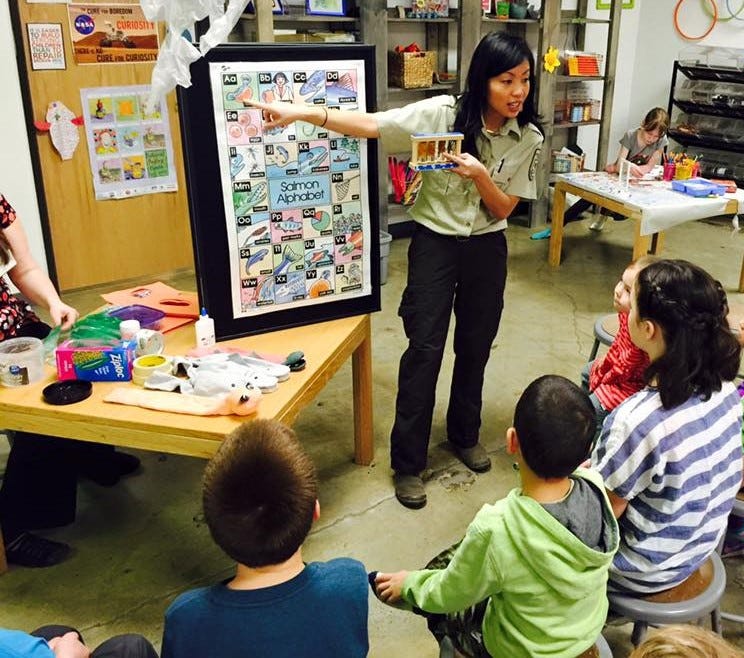 A woman in a brown uniform shows a group of children a graphic about salmon.