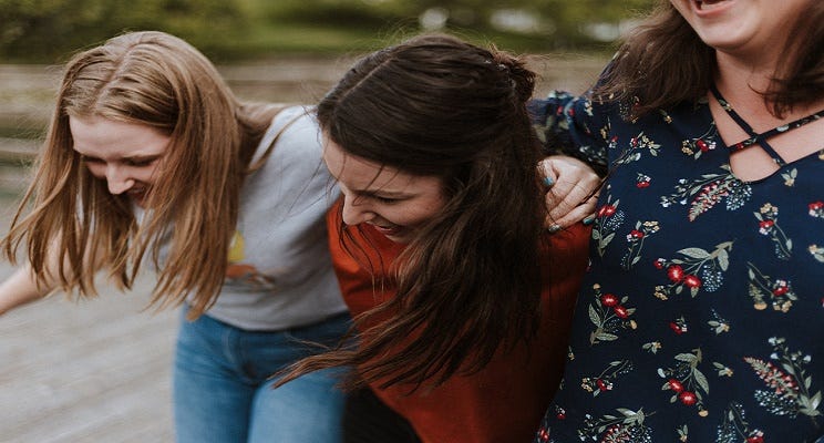 Three laughing women walk side by side with their hands on each other’s shoulder.