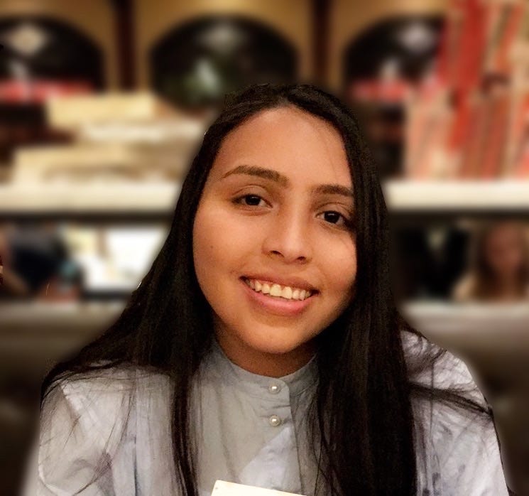 A young woman smiling in front of a blurry background. You can see her face and shoulders only. She is wearing a grey shirt.