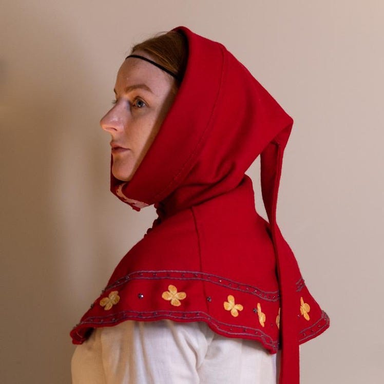 A red medieval knit hood that drapes as a collar around the shoulders with yellow four-point flowers on the border. It is being worn by a white female looking to the side against a plain white backdrop.
