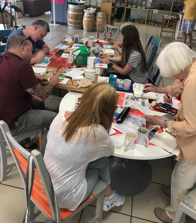 A group of people sit at tables covered with craft supplies. They are painting and drawing.