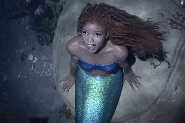 A young Black woman with red locs, actress Halle Bailey, sits on a rock underwater. She is wearing a purple bustier and has a shimmering green mermaid tail. She is looking up, and her mouth is open in song.