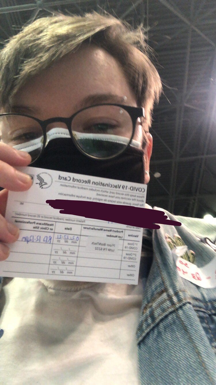 A picture of the author holding up his vaccination card. He is wearing two masks, glasses, and is in the Javits Convention Center. His personal information has been blocked out of the photo.