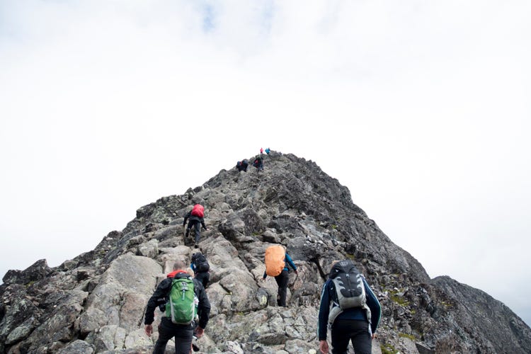 Climbers, backs to the viewer, hiking their way up the side of a rocky mountaintop in an effort to reach the summit