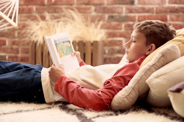 Boy on a bed, propped up by pillows, reading a book.