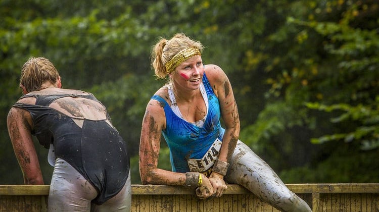 Two women covered of dirt on exercise atire are going over a fence. One of the has her face to the camera and looks tired.