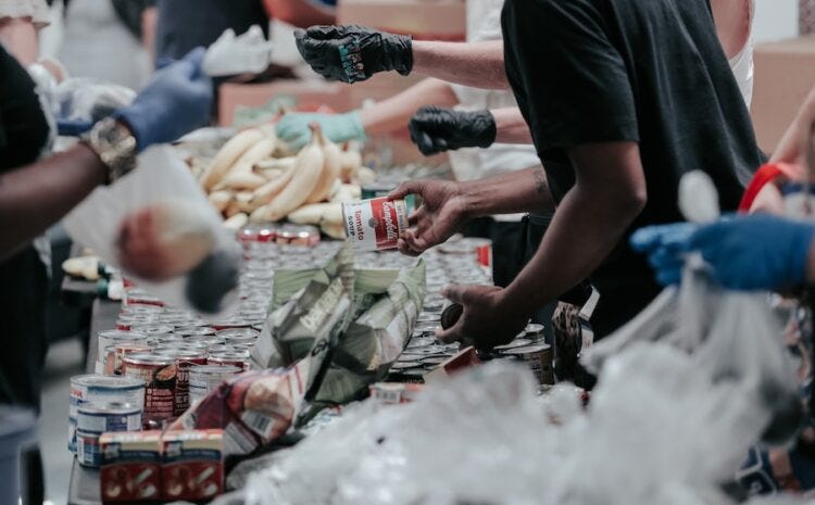 Photograph of a food bank with a table full of canned goods — several people are passing food back and forth across the table. Their faces are not visible