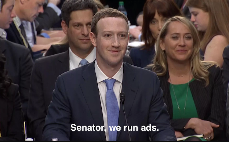 Screenshot of Mark Zuckerberg during 2018 hearing at the US Senate following Cambridge Analytica data breach. Mark Zuckerberg is sitting at foreground, wearing a suit. Behing him, a female and a male are sat, also wearing suits. There are a lot of unidentified people all sat behind them. The three of them are having a smug smile, due to Mark Zuckerberg answer to Sen. Orrin Hatch asking how Facebook was sustaining itself while being free for users. Mark Zuckerberg answered “Senator, we run ads”