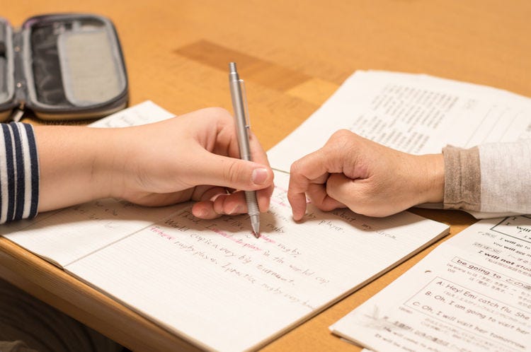 Schoolwork on a desk, a hand over it holding a writing implement, and a second hand seemingly pointing out something on the page.