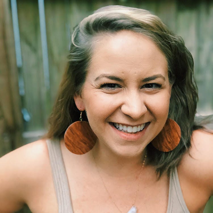 A portrait of Jess Budnick. She is a white woman smiling with brunette wavy hair and large brown wood circle earrings.