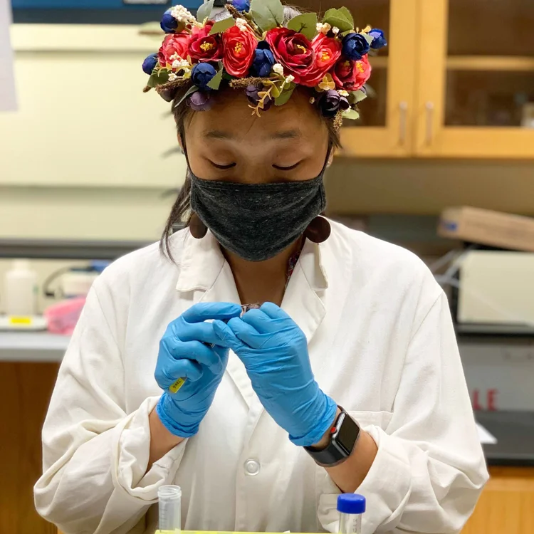 A women wearing a black face mask, white lab coat and blue rubber gloves. She is holding a tube and working on an experiment in a science lab. She also has a really colorful flower tiara on her head.