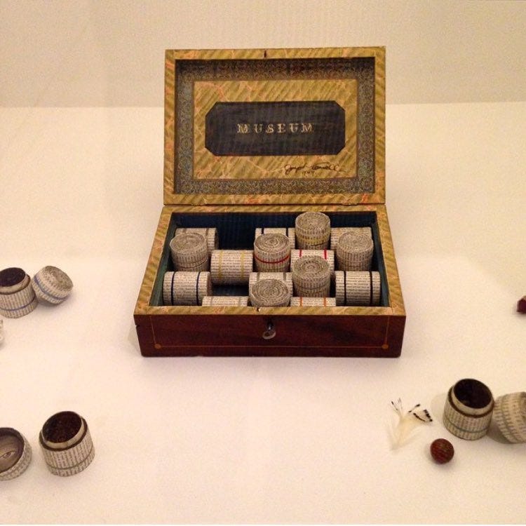 A small wooden box with its lid open. Inside the lid is a label that simply says “MUSEUM”. Inside the box are a number of small cylinders which look to be made out of either thing wood or cardboard. On the shelf outside the box, several similar cylinders are open with their contents viewable, featuring small objects like paper clips, string, etc.