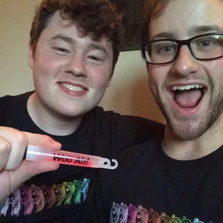 A photo of the author and a friend wearing rainbow Kim Petras tee shirts and holding up a “Woo Ah!” glowstick.