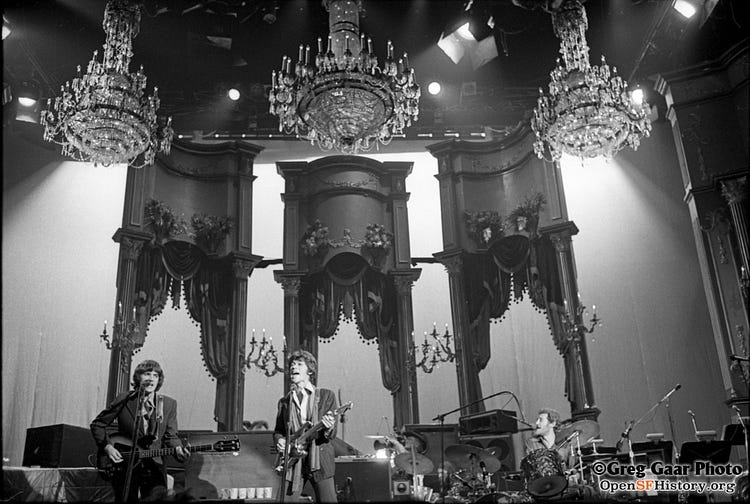 Black and white photograph of The Band playing on the Winterland stage under chandeliers. 