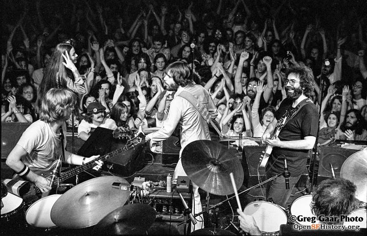 Black and white photo of The Grateful Dead on stage with an audience of fans in the background.