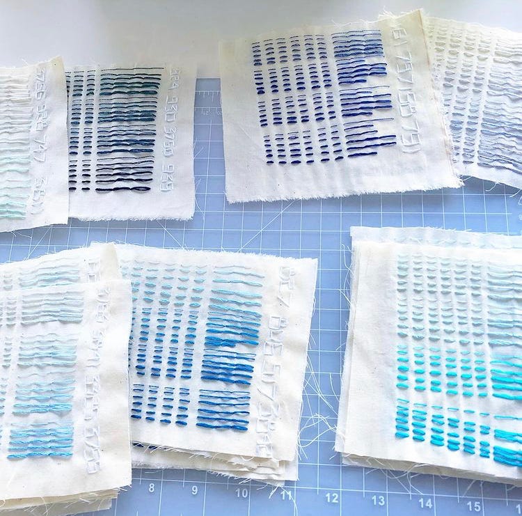 Fabric pages scattered across a light grey cutting mat. The pages show stitches in many shades of blue, from light sky blue and bright teal, to deep ocean and dark navy. Each color is stitched in sections with six rows. Each row shows simple stitches in a progression of thickness, from one to six threads.