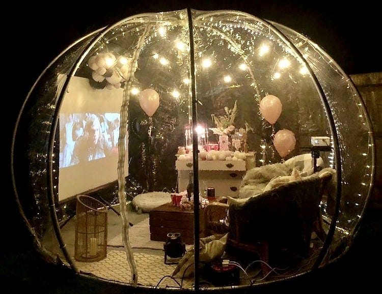 Image of the outdoor party pod at night, complete with cinema kit, fairy lights, big comfy armchairs and pink balloons.