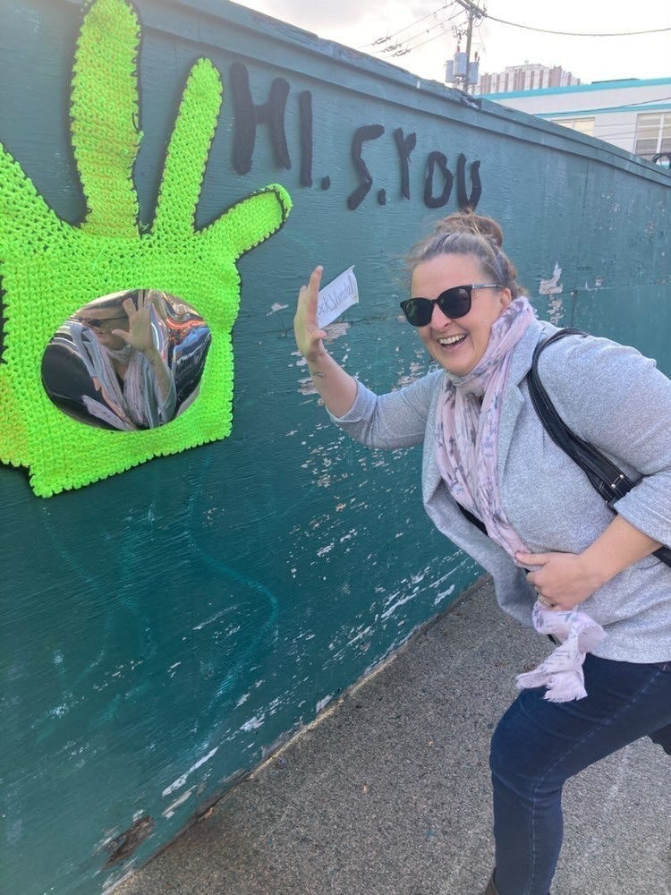 A smiling blonde woman with a top knot and sunglasses wearing a grey sweater and pink scarf holds her hand in a “High Five” gesture in front of a neon green crocheted hand with a mirror in the centre.