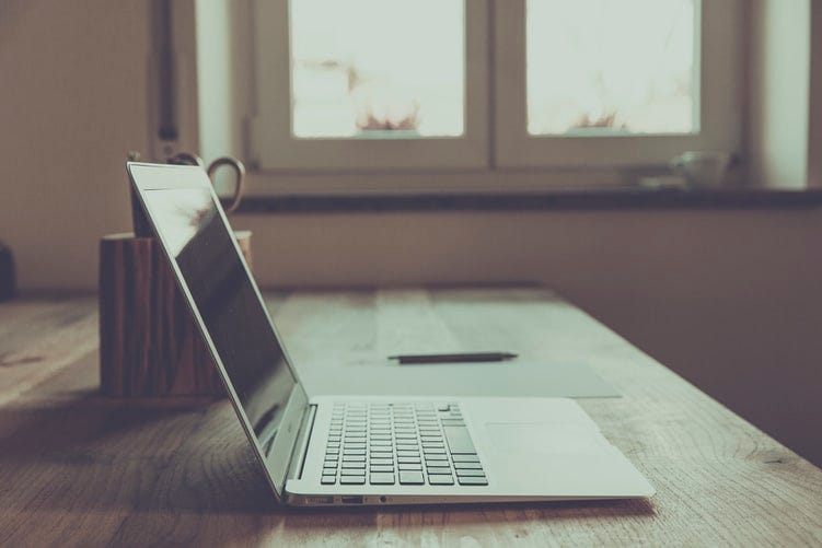 An open white laptop on a brown wooden table. A pen and mug rest on the table beside it. A while wall and closed window are in the background, illuminating the room with daylight.