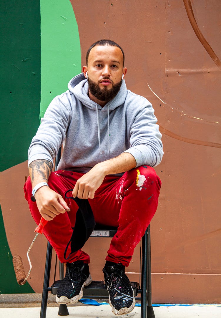 Alexandre Keto sitting on a stool in front of his mural while holding a paint roller.