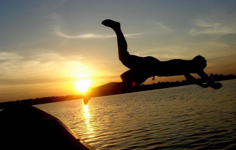Man jumping off of boat into lake with sunset in background.