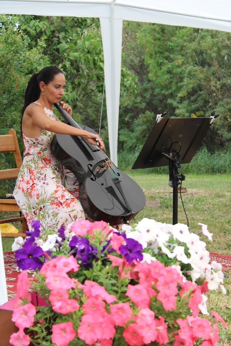 Woman in light dress sits playing electric cello behind flowers