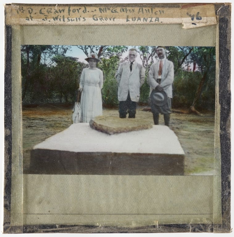 Lantern slide featuring image of one white woman and two white men. They are dressed in early twentieth century western attire and are stood behind a large flat tombstone.