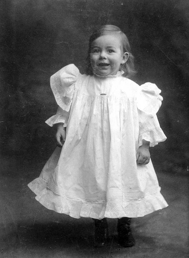 Baby Hemingway stands smiling in a dress with puffy sleeves.