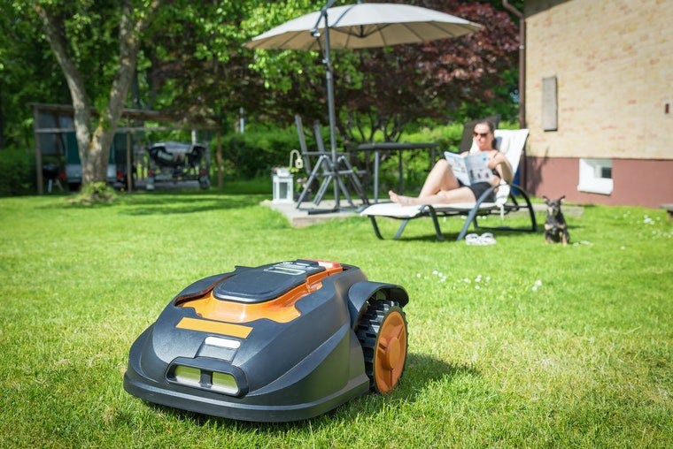 A robotic lawnmower mows the grass while in the background a person reads in a lounge chair.