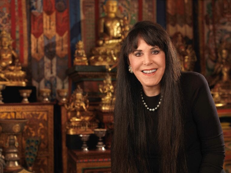 A portrait of a woman with long brown hair stands before a Buddhist shrine of glittering statues and paintings.