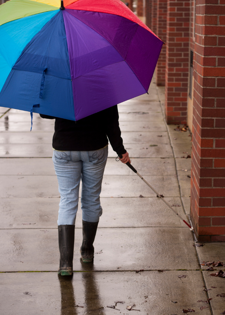 Fotografía de una persona camninando con un bastón blanco en la mano derecha. En la mano izquierda lleva un paraguas con los colores del arcoiris. Crédito: Canva