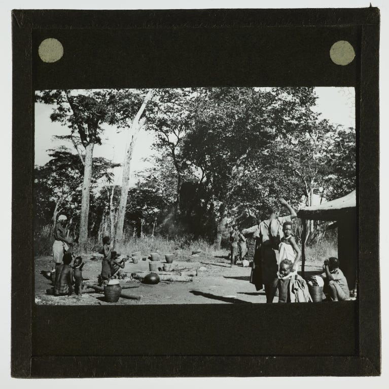 Black and white image of an African village with indigenous men, women and children. There is a building on the right and trees in the background, and some large pots and baskets on the ground.