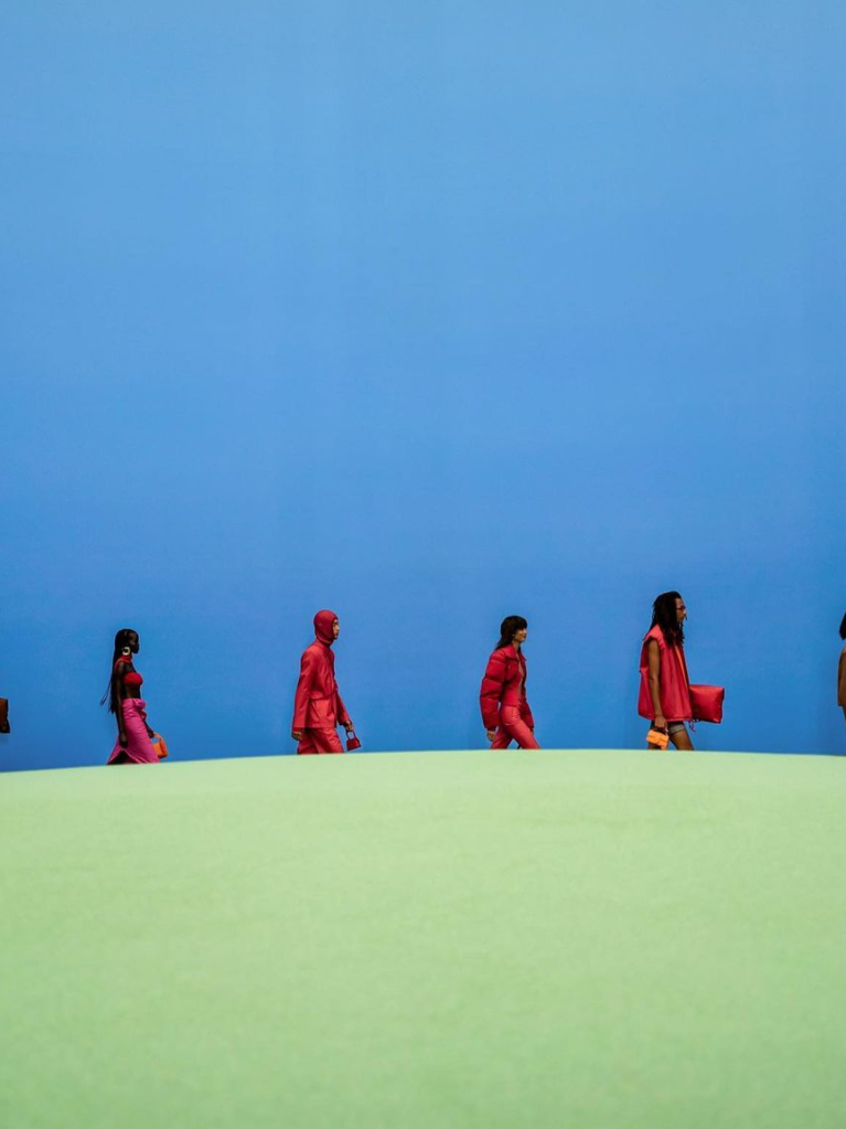 Models wearing red outfits walking across a green hill in a line with a blue backdrop.