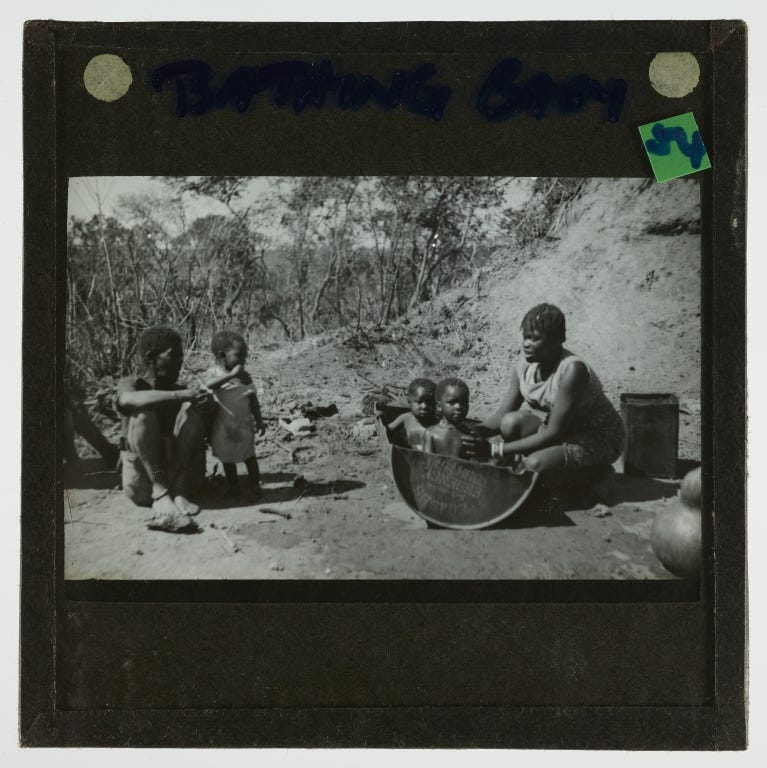 Black and white photograph featuring a woman crouched on the right of the image bathing two children in a tin bath. A man is sitting on the ground and a third child is standing next to him. All of the people featured are black African.