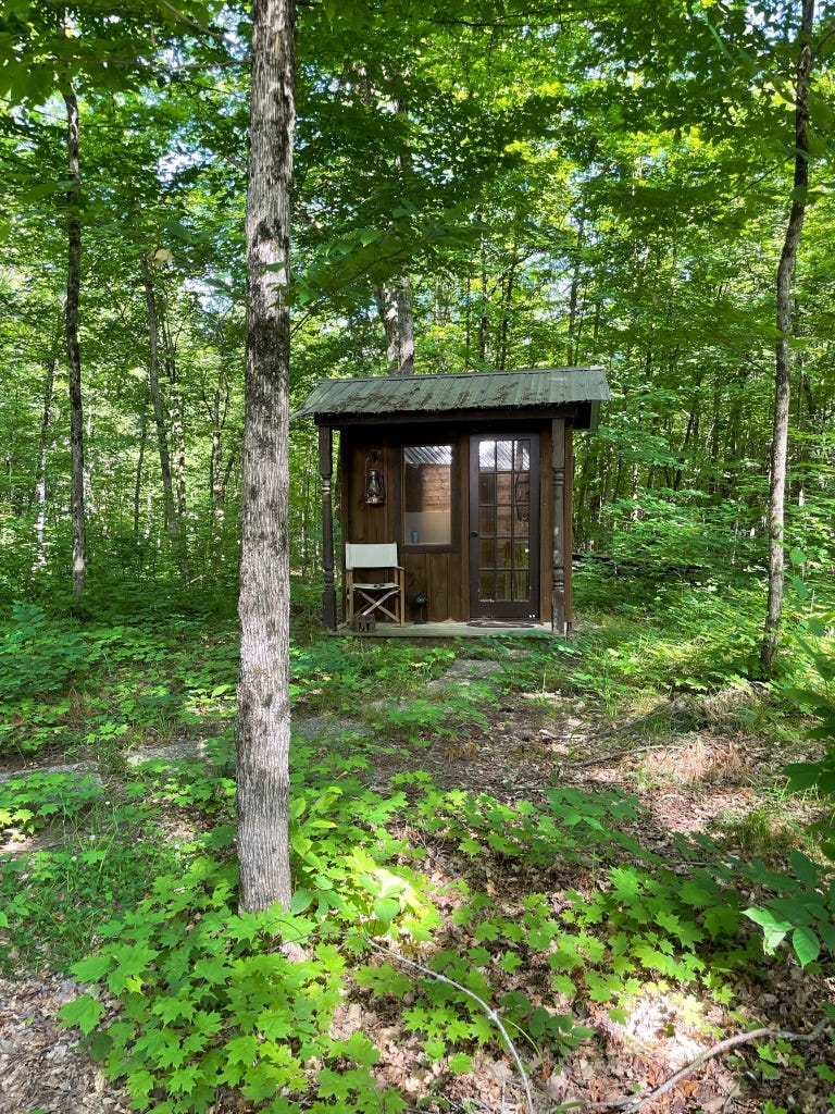 A private gem of an outhouse with a porch, window, and glass-paned door sits in the woods behind Bijou Glabin.