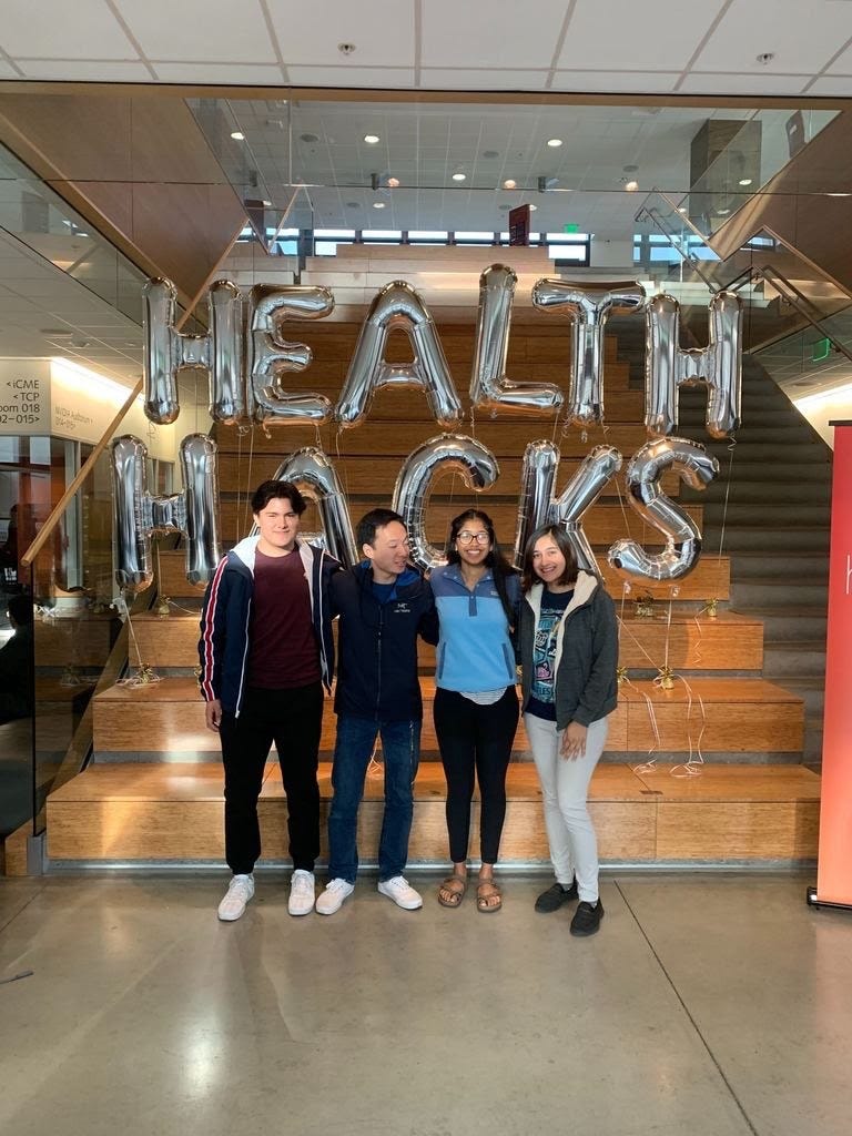 4 young adults pose in front of balloons that read “Health Hacks.”