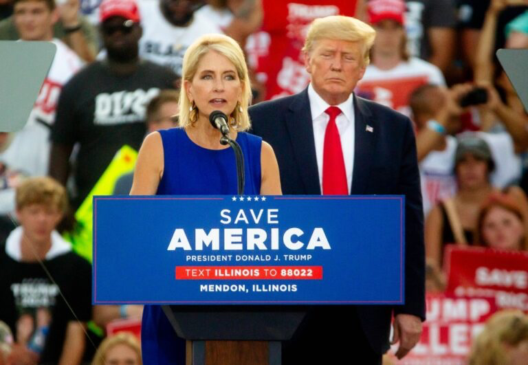 Rep Mary Miller, wearing a dark blue dress, stands at a podium and speaks to a crowd at a “Save America!” Rally. Trump stands slightly behind her to the right, looking down towards the podium, arms at his side, his expression somewhat pinched and vacant. A crowd sits and stands in the risers behind them holding signs of various sizes and colors.