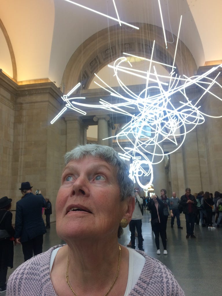 A white woman in her eighties with short grey hair looks up at the Tate Britain Gallery in London. Above her head is a light sculpture that makes her appear to have all kinds of inspiration.