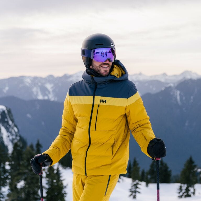 Man on top of the mountain wearing a yellow and black color block Helly Hansen ski Jacket about to ski down hill.