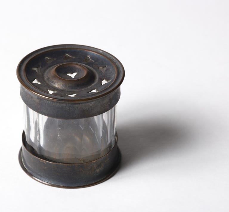 Sand shaker belonging to Elisabeth Gaskell, the body is made of colourless glass and the top and bottom are made of metal. There are small heart shaped holes at the top where the sand can be shaken out on to a handwritten letter to help dry the ink.