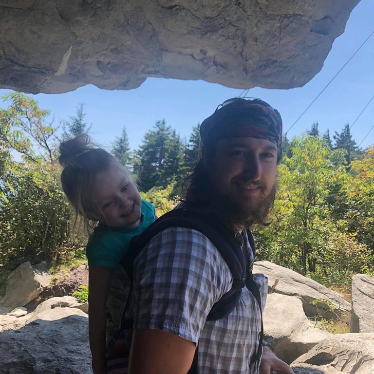 A picture of Mitch carrying Josie in a baby carrier with a woods in the background.