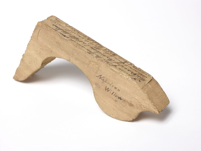 Curio from Isabella Banks collection; a small piece of wood from the Willow tree which grew over the grave of Napoleon I at St. Helena. It was cut down when the body was exhumed and removed to Paris. A slip of paper with handwritten text describing the curio has been attached to the upper edge of the wood.