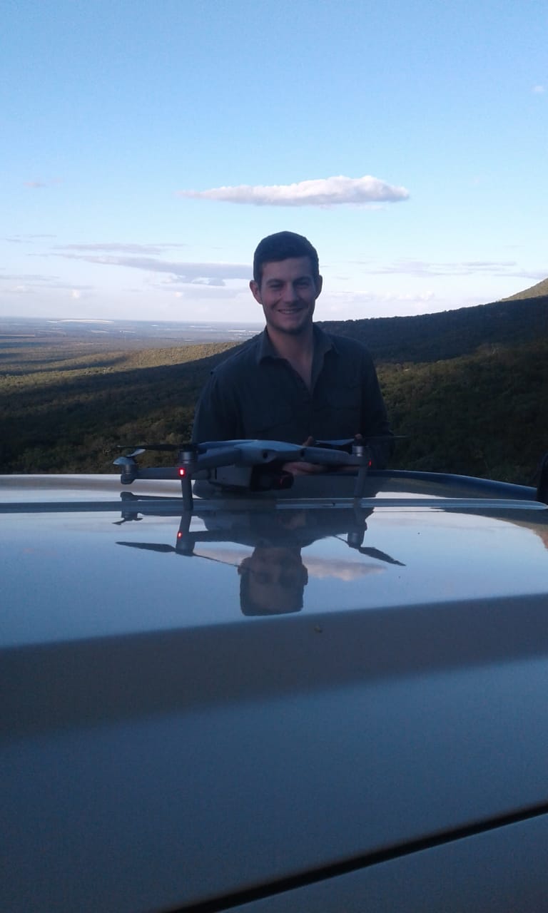 A Caucasian male smiling, with a drone on the bonnet of a car in front of him, and a mountain in the background