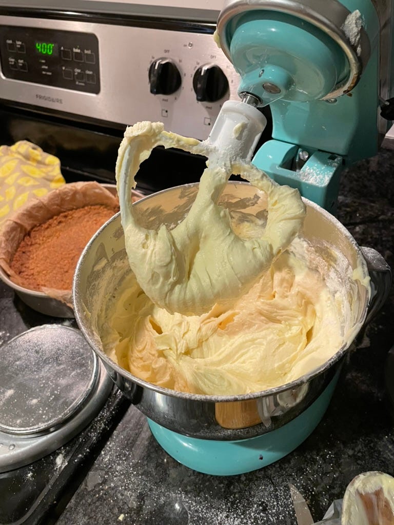 A stand mixer full of cheesecake batter, with a graham cracker crust next to it on top of a stove.