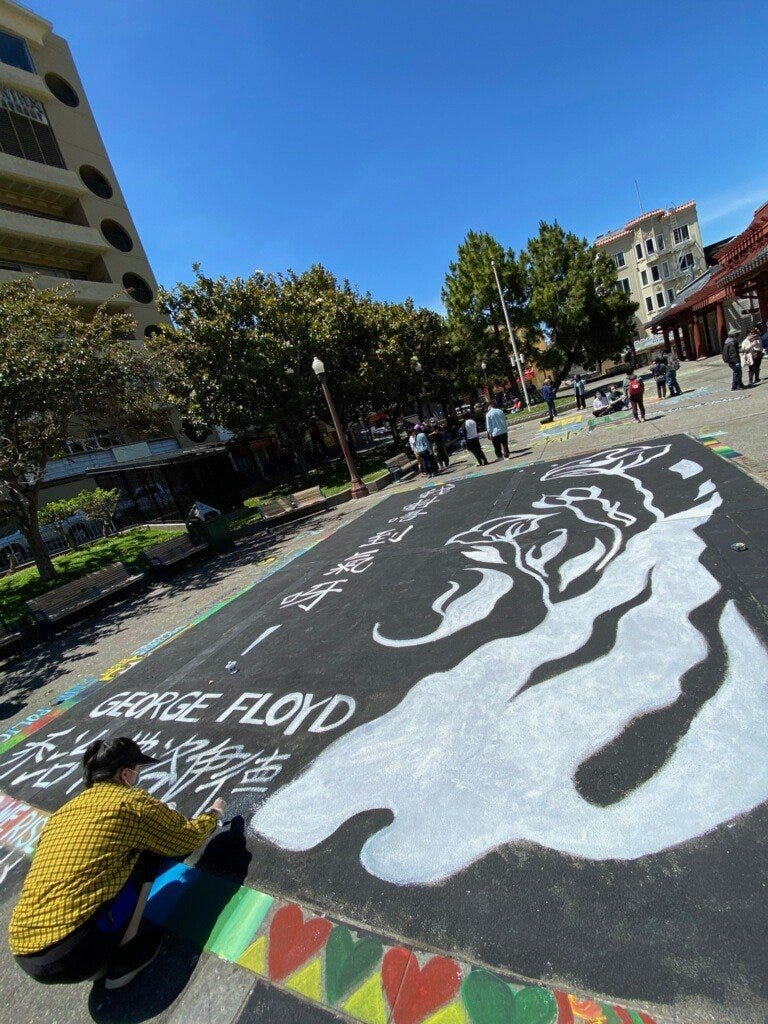 A young Asian woman kneels on the asphalt in a park, painting a mural that says “George Floyd” and Chinese characters.