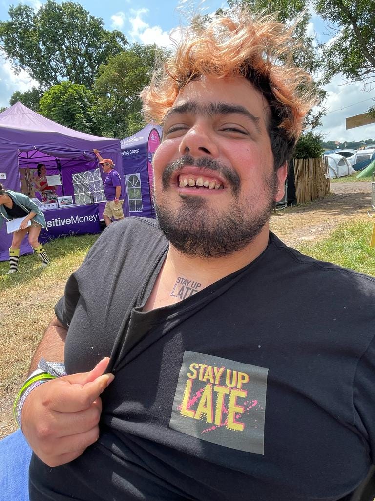 Man with a learning disability at Glastonbury Festival wearing a black t-shirt with Stay Up Late on it and a temporary tattoo saying Stay Up Late on his neck.