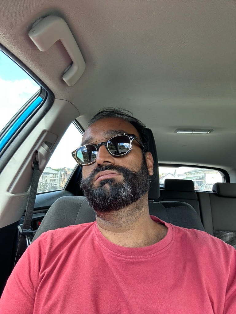 Selfie of brown man in car. Leaning back and looking tired. Beard and sunglasses. Red t-shirt.